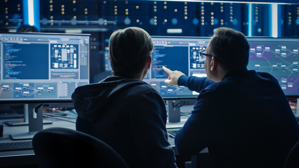 Dos ingenieros observan pantallas con esquemas y código de arquitectura de datos en un entorno corporativo.