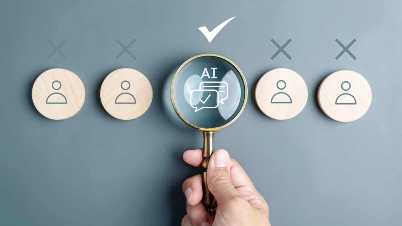 Imagen de una mano sosteniendo una lupa sobre un token central que dice "AI" con burbujas de diálogo y una marca de verificación. Otros cuatro tokens con iconos de usuario tienen una 'X' encima de ellos, simbolizando la AI como la opción elegida frente al talento humano tradicional en los despidos tecnológicos de 2026.