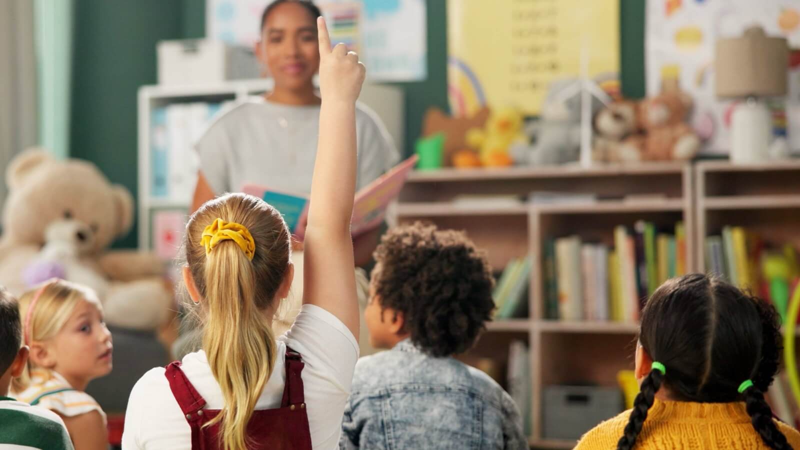 Un grupo de niños de primaria sentados en el suelo de un salón de clases, mirando hacia una maestra que sostiene un libro. Una niña en primer plano levanta la mano para responder una pregunta. Fondo con estanterías de libros y juguetes. Fotografía de stock.