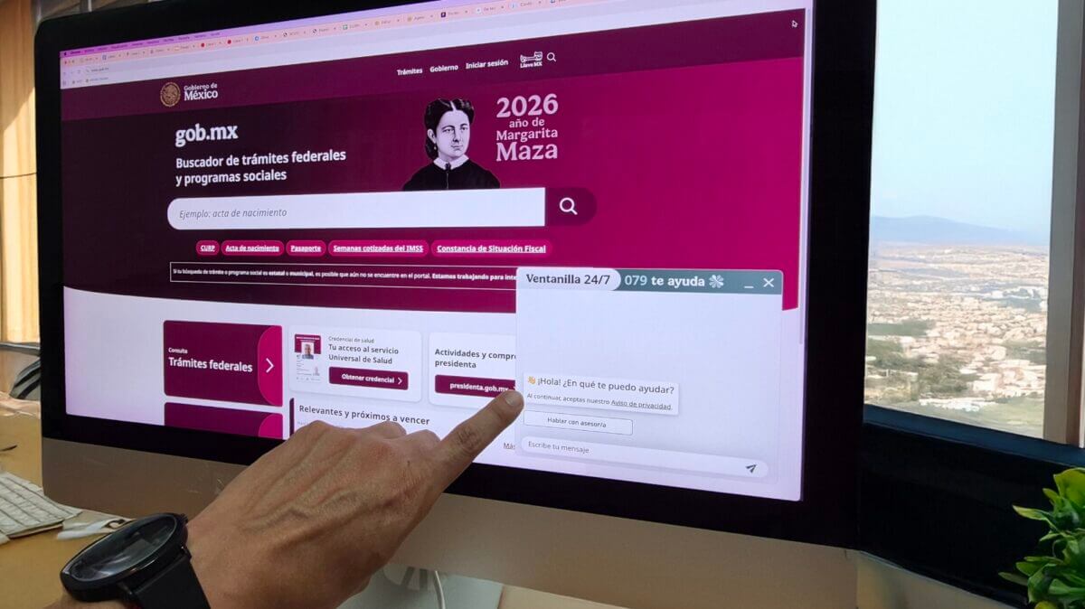Fotografía de primer plano de una mano interactuando con la interfaz de "Ventanilla 24/7", un chatbot de IA generativa (GenAI) desarrollado por la ATDT, en el portal oficial gob.mx en una pantalla de escritorio. La pantalla muestra el buscador de trámites federales y el widget de chat abierto.