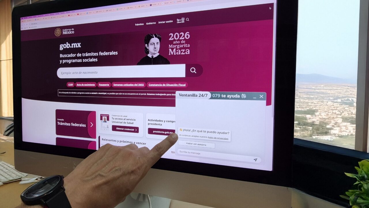 Fotografía de primer plano de una mano interactuando con la interfaz de "Ventanilla 24/7", un chatbot de IA generativa (GenAI) desarrollado por la ATDT, en el portal oficial gob.mx en una pantalla de escritorio. La pantalla muestra el buscador de trámites federales y el widget de chat abierto.
