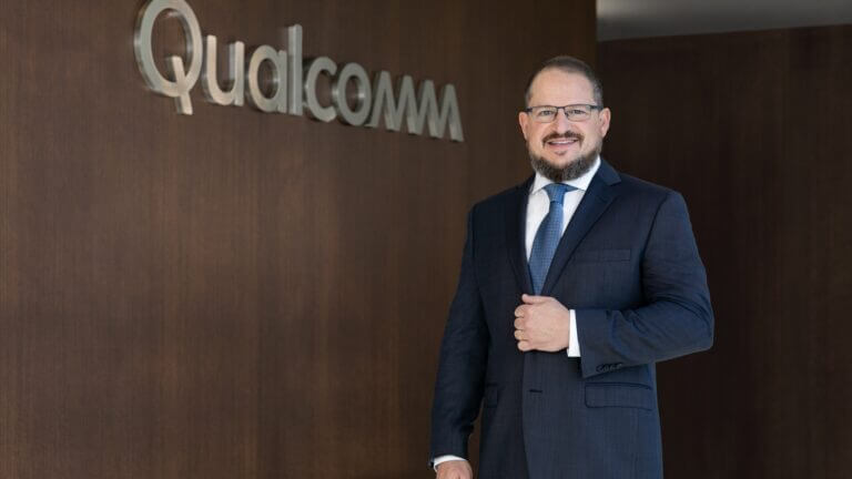 Cristiano Amon, CEO Qualcomm AI agéntica, posa en traje frente al logo metálico de Qualcomm en pared de madera.
