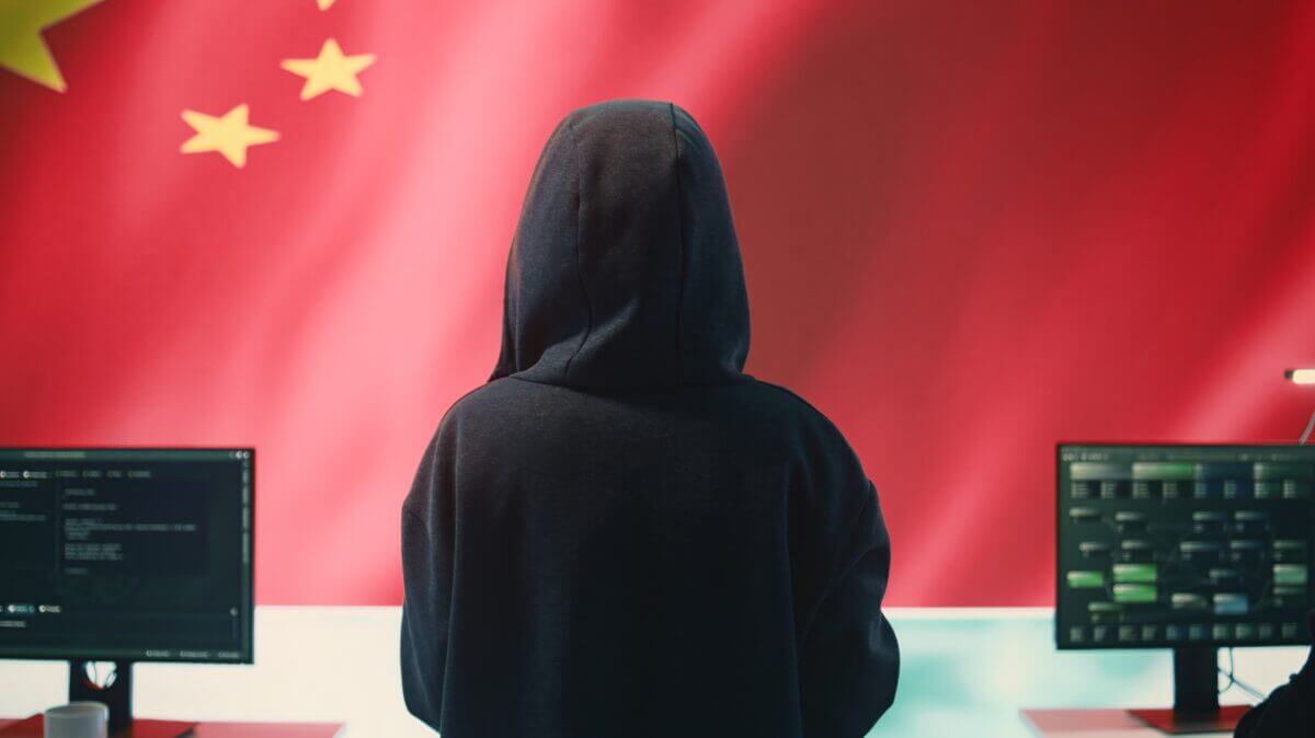 Un hacker o espía informático de espaldas con capucha frente a monitores de computadora, con la bandera de China de fondo, ilustrando los riesgos de espionaje de China en LinkedIn.