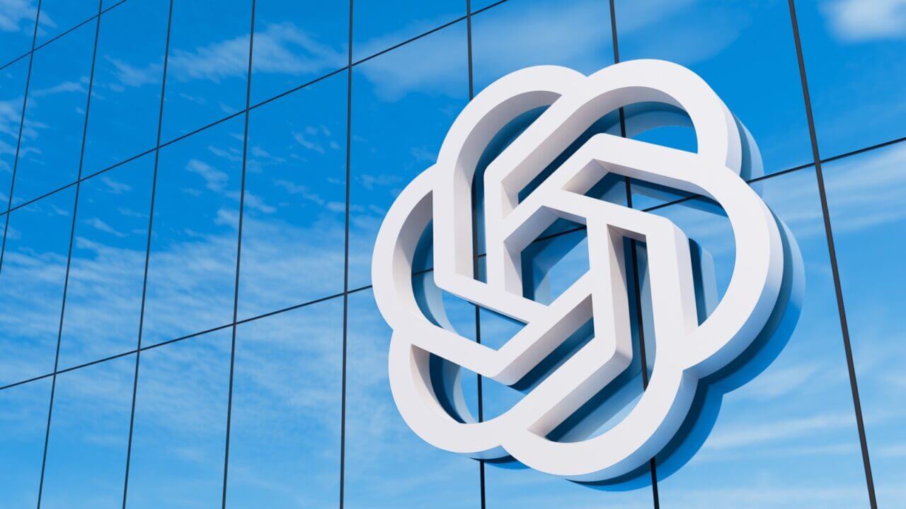 El logotipo de OpenAI, una estructura geométrica entrelazada blanca y hexagonal, montado en la fachada de un rascacielos de vidrio azul moderno con reflejos de nubes.