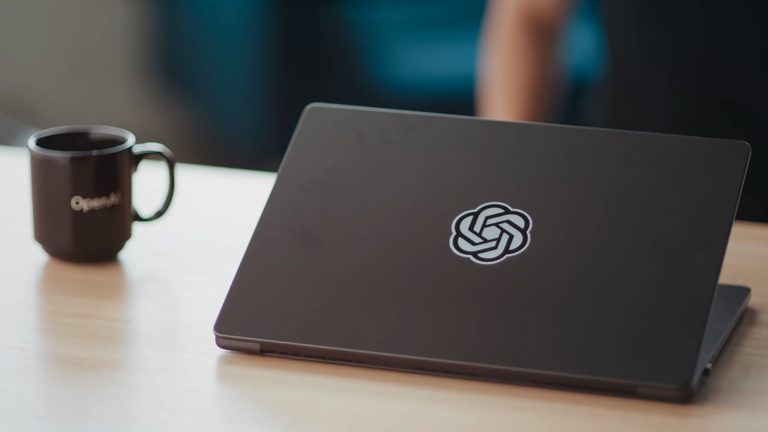 Laptop con el logo de OpenAI junto a una taza de café en un escritorio, ilustrando los costos de tokens OpenAI.